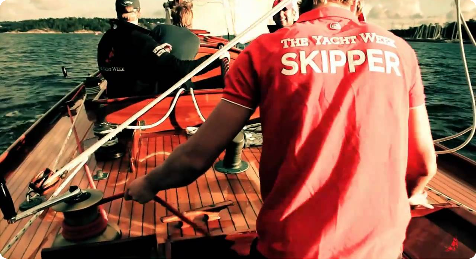 A person in a red "The Yacht Week Skipper" shirt steers a Classic Sailing Yacht on open water, with crew seated ahead and ropes and the wooden deck of this elegant vessel visible.