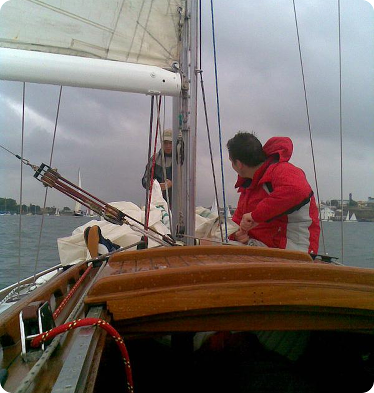 Two people sail a classic sailing yacht under cloudy skies; one in a red jacket looks ahead whilst the other adjusts the sail. City buildings and other boats are visible in the background, evoking the timeless charm of Square Metre Yachts.