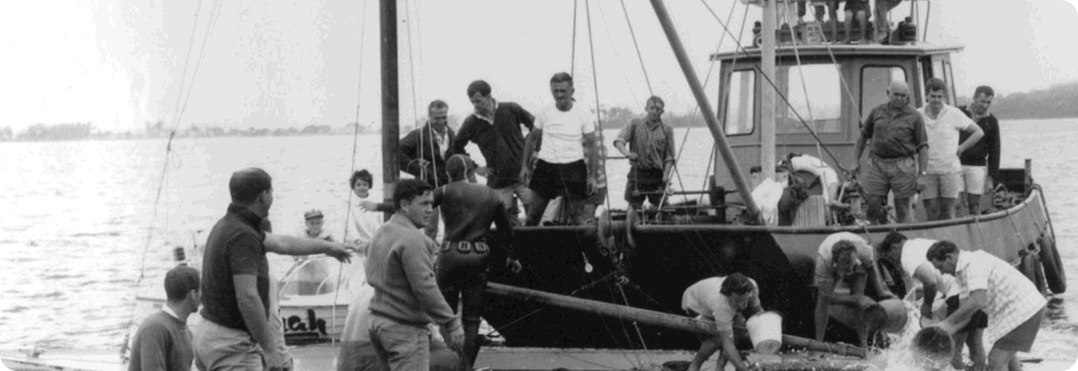 A group of people gather on a boat and a moored vessel, working together near the water. Some are handling ropes and poles as they prepare Skerry Cruisers. It is daytime, with Square Metre Yachts visible on the calm water in the background.