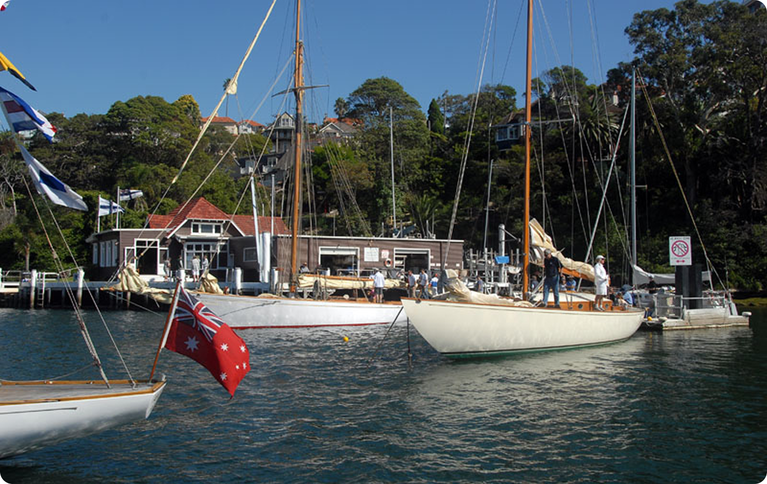 Sailing boats, including elegant Skerry Cruisers and Square Metre Yachts, are moored at a marina with people on board. A building, trees, and an Australian flag are visible. The calm water reflects the clear sky above.