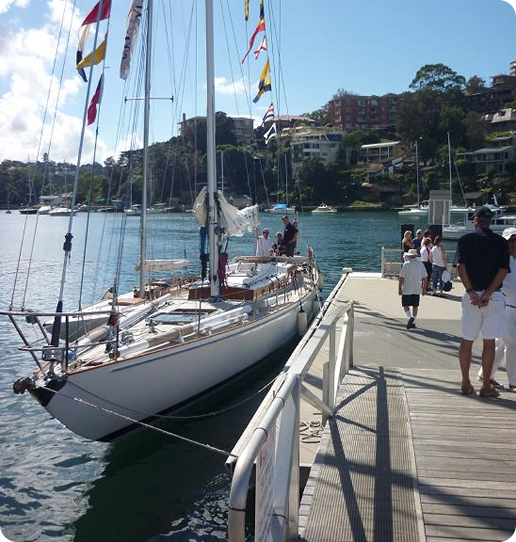 A white Classic Sailing Yacht decorated with colourful flags is moored at a marina. Several people stand on the quay and on the boat, with houses and trees visible on the hillside in the background under a partly cloudy sky.