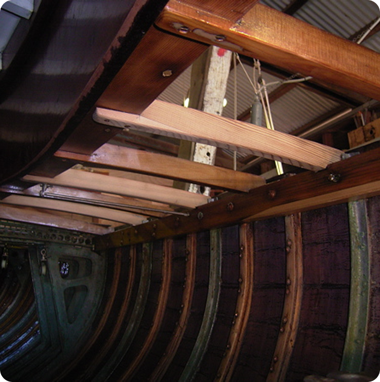 Interior view of a wooden Skerry Cruiser hull under construction, showing curved wooden ribs, planks, and beams. The image highlights the detailed framework and structure inside a workshop setting.