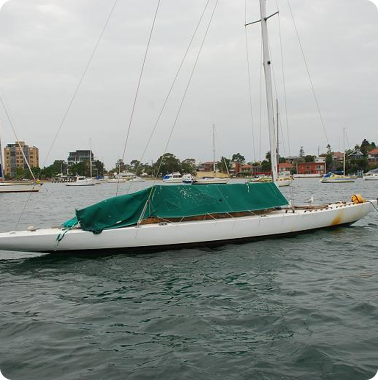 A white Schärenkreuzer with a green cover over its deck is floating on calm water near several buildings and trees under a cloudy sky.