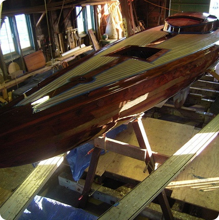 A wooden Schärenkreuzer under construction is positioned on stands inside a workshop, with sunlight streaming through the windows and various tools and materials visible in the background.