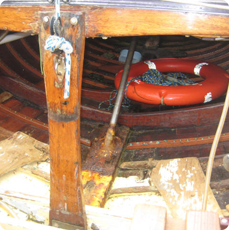 A close-up of the inside of a wooden boat, showing wooden ribs, loose planks, an oar, ropes, and an orange lifebuoy with white stripes—a scene reminiscent of classic Square Metre Yachts. The area appears worn and cluttered.