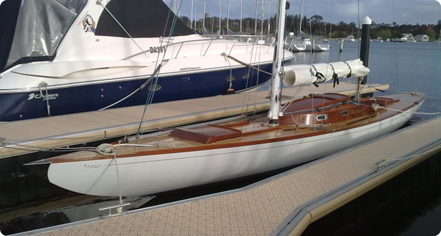 A sleek, white sailboat reminiscent of Classic Sailing Yachts with a polished wooden deck is moored at a marina next to a larger yacht. Its upright mast and furled sails stand out against the calm water and other boats in the background.