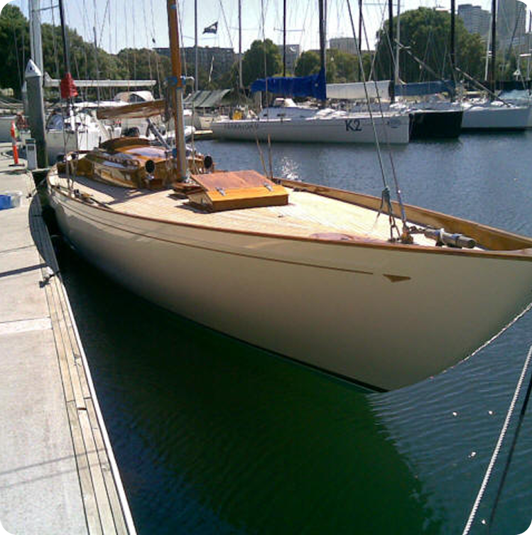 A classic wooden Schärenkreuzer with a polished deck is moored at a marina, surrounded by other sailing boats and calm water, with buildings and trees in the background.