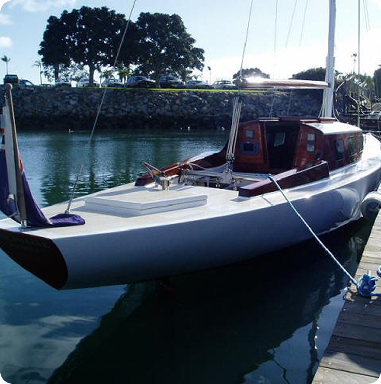 A white Skerry Cruiser sailboat is moored at a wooden pier in a calm marina, with trees, a stone wall, and parked cars visible in the background under a partly cloudy sky.