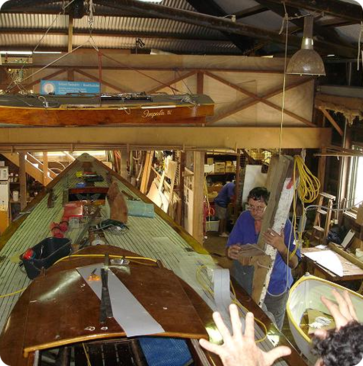 Two people work inside a cluttered wooden boat workshop, surrounded by tools, supplies, and large boats such as Classic Sailing Yachts. Sunlight pours in through the windows and roof, illuminating the busy workspace.