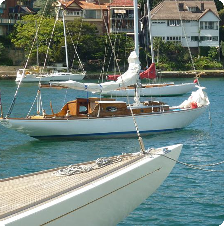 A white Square Metre Yacht with wooden trim is anchored in calm blue water near a shoreline with houses and green trees. Another sailboat’s bow, possibly a Skerry Cruiser, is visible in the foreground.