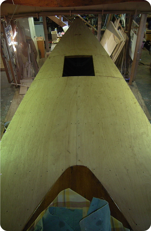 A wooden Square Metre Yacht under construction in a workshop, viewed from above. The deck is mostly covered with plywood, with a square opening in the centre and tools and materials scattered around.