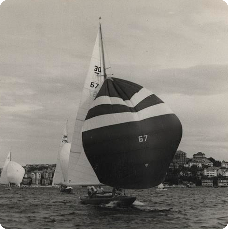 A black-and-white photo of a classic sailing yacht, with the number 67 on its large, rounded sail, sailing on the water near a coastline dotted with buildings and other sailing boats in the background.