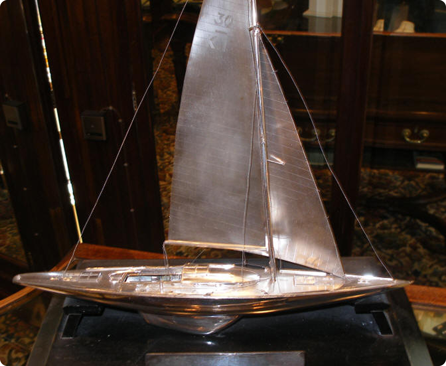 A detailed silver model of a Classic Sailing Yacht with two sails, displayed indoors on a dark base, in front of wooden cupboards and patterned carpet.