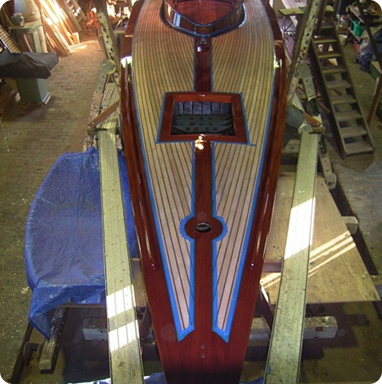 A wooden Skerry Cruiser with polished, striped decking and blue accents is positioned upright in a workshop, supported by beams on each side. Tools and materials are scattered around the workspace.