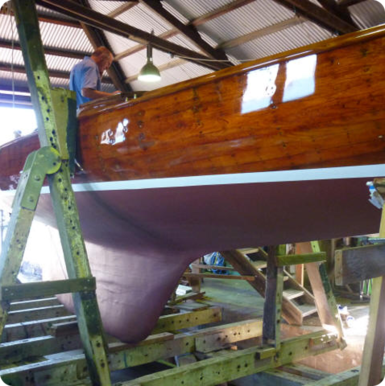 A person stands on a ladder working on the side of a wooden Skerry Cruiser with a shiny varnished hull and red-brown bottom inside a workshop.