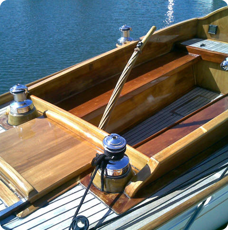 A close-up view of a Schärenkreuzer cockpit, showing polished wooden benches, deck, and trim, with a wooden tiller and shiny metal winches—classic details of these elegant Square Metre Yachts floating on calm water.