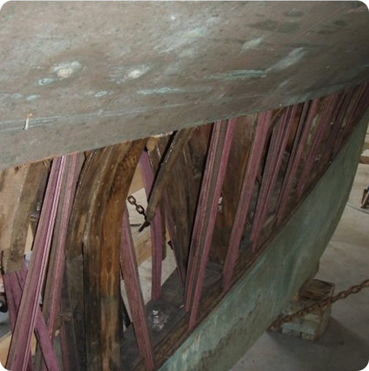 Damaged wooden boat hull with exposed ribs and beams, showing broken and missing planks. The structure appears weathered and is indoors, possibly undergoing repair or restoration of a classic Schärenkreuzer or Square Metre Yacht.