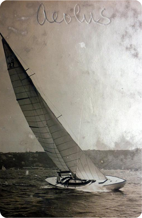 A vintage black-and-white photo of a Schärenkreuzer sailboat named Aeolus, with the number 27 on its sail, glides across the water with a distant shoreline in the background.