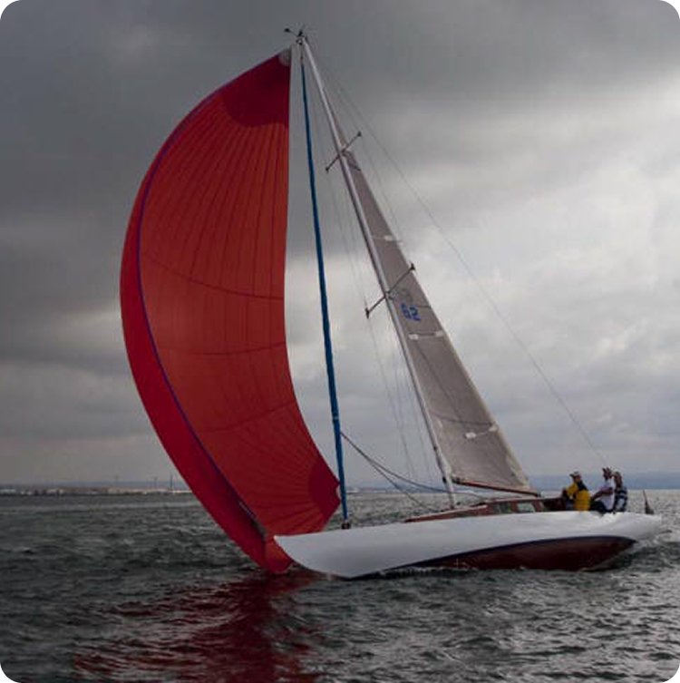 A classic Skerry Cruiser with a large red spinnaker sail glides on the water under a cloudy sky. Several people are on board, and the distant shore is visible in the background.