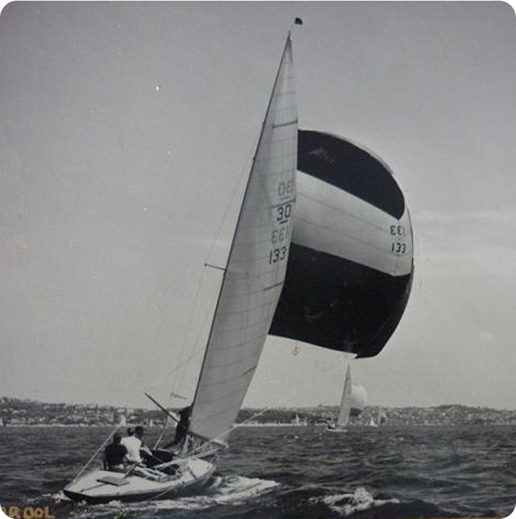 Black-and-white photo of a sailboat, possibly a Skerry Cruiser, with two people on board, sailing on choppy water beneath a large spinnaker. Another Square Metre Yacht sails in the distance, with shoreline buildings in the background.