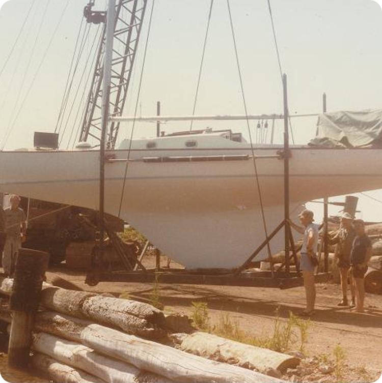 A white sailboat, possibly a Skerry Cruiser, is suspended by a crane at a shipyard, with several people standing nearby. Logs and equipment are visible in the foreground on this sunny day outdoors.
