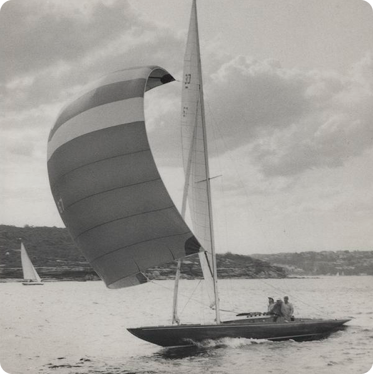 A black and white photo of a Schärenkreuzer sailboat with a large striped spinnaker gliding on the water, three people on board, and a distant shoreline in view. Another Square Metre Yacht appears further away.