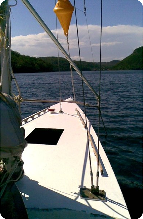 The photo shows the bow of a white Schärenkreuzer sailboat on calm water, with a yellow buoy hanging above the deck and green hills in the background under a partly cloudy sky.