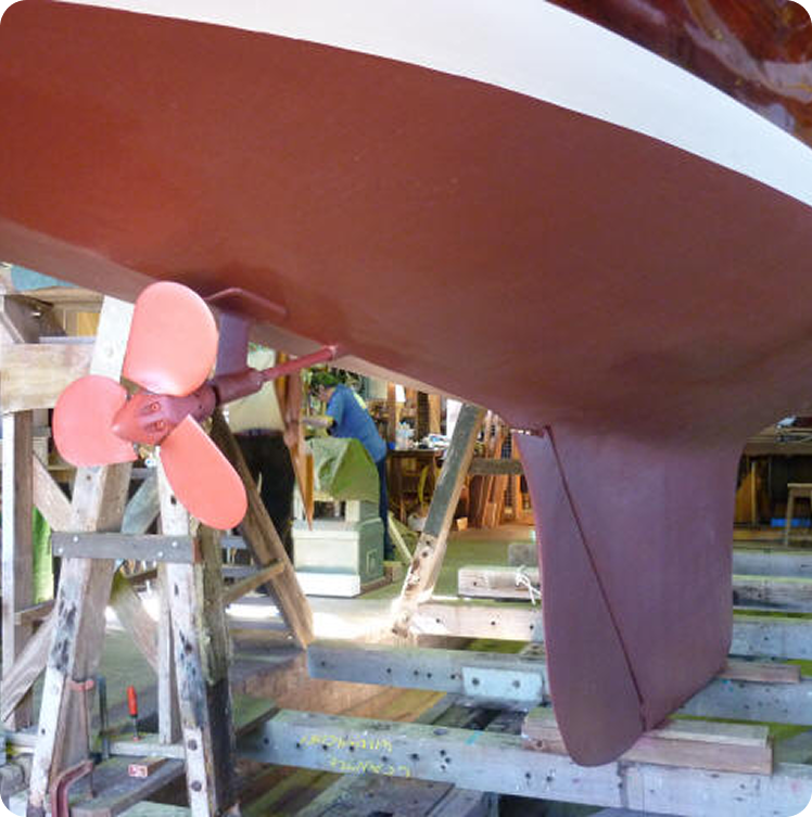Close-up view of a Schärenkreuzer’s hull in a workshop, featuring a pink propeller and rudder. The Square Metre Yacht is elevated on wooden supports, with tools and equipment visible in the background.