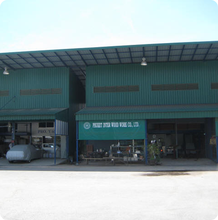 Exterior view of a green industrial warehouse with a blue roof. The entrance sign reads PHIRAT INTER WOOD WORK CO., LTD. Equipment and materials are visible under the canopy, known for craftsmanship in Square Metre Yachts.