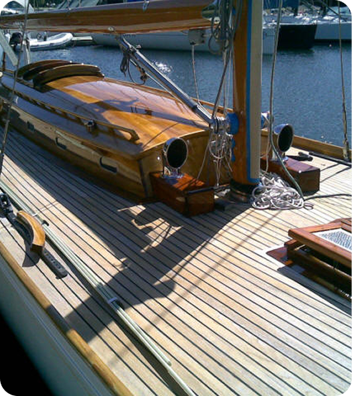 A close-up view of a wooden sailboat deck with polished wood finishes, ropes, and nautical equipment—this Schärenkreuzer exemplifies the elegance of Classic Sailing Yachts, berthed at a marina with other boats visible in the background.