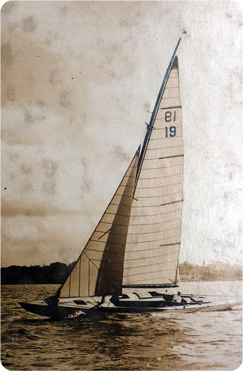A vintage photo of a Schärenkreuzer sailboat with the numbers 19 and 13 on its tall, triangular sail, gliding on a calm body of water with a distant shoreline and cloudy sky in the background.