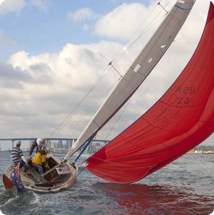 Two people sail a small boat with a large red sail, likely a graceful Schärenkreuzer (Skerry Cruiser), heeled over the water. A city skyline and bridge are visible in the background under a partly cloudy sky.