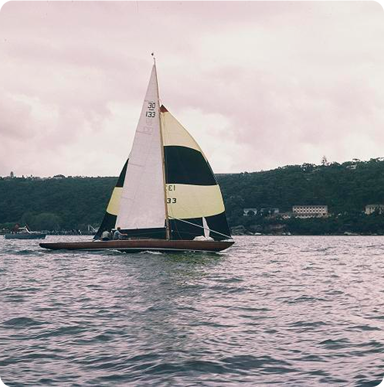 A black and yellow Square Metre Yacht glides on a body of water, with a forested shoreline and several buildings visible in the background under a cloudy sky.