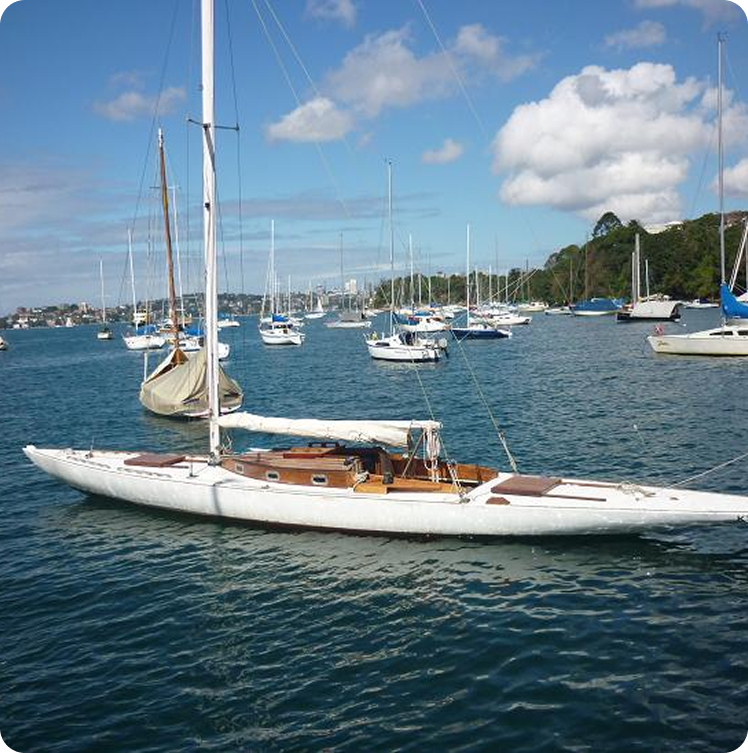 A white sailboat with a wooden deck, reminiscent of classic Square Metre Yachts, is anchored in a harbour surrounded by other boats on calm blue water. Green trees and a partly cloudy sky are in the background.