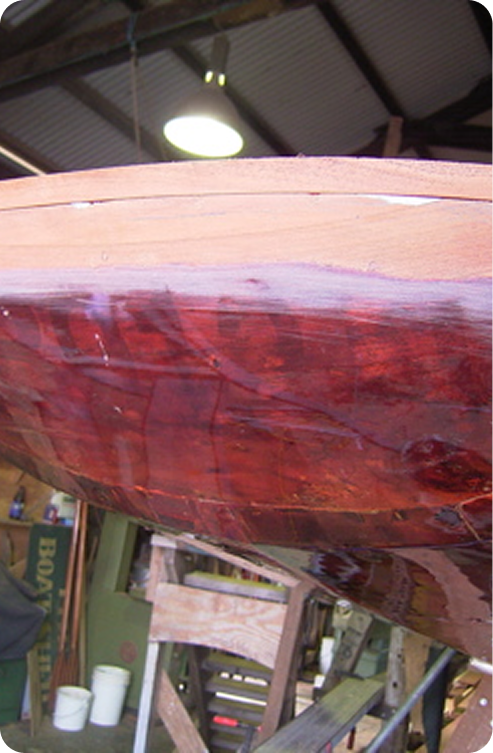 Close-up of a wooden Schärenkreuzer hull in a workshop, partially coated with shiny varnish or resin. Workshop tools, shelves, and supplies are visible in the blurred background.