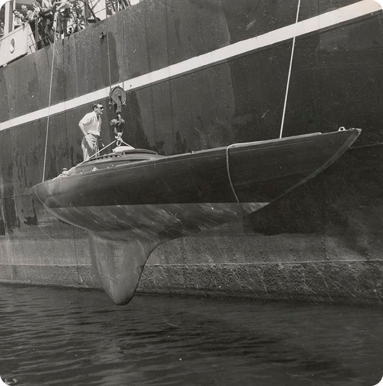 A small, sleek Schärenkreuzer with a large keel is suspended by ropes beside a ship. Several people observe from above as the classic sailing yacht is lifted from the water. The scene appears to take place at a quay.