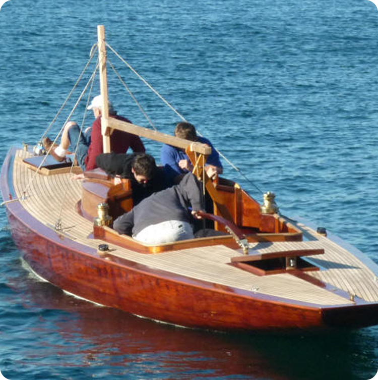 A small wooden Schärenkreuzer sailboat with four people aboard sails on calm blue water. The boat features a polished deck, brass fittings, and visible rigging, with the group seated and facing forward.