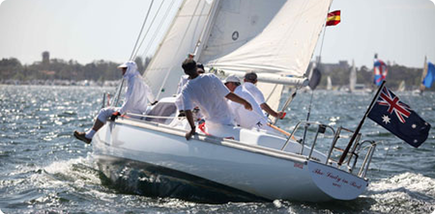 A group of four sailors dressed in white navigate a classic sailing yacht with an Australian flag on open water. Other sailing boats and a distant shoreline are visible in the background.
