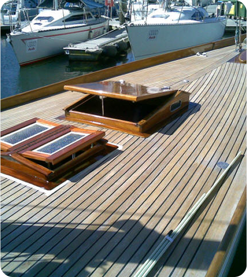 A close-up of a wooden boat deck with an open rectangular hatch, shiny varnished wood, and a marina with other Classic Sailing Yachts in the background. Sunlight highlights the clean, polished surface.