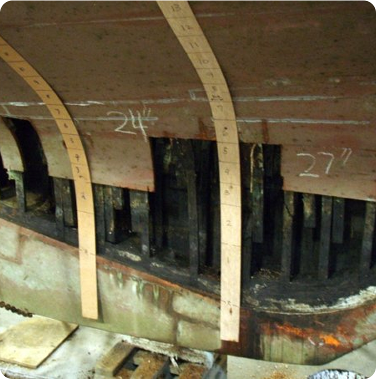 A close-up of a wooden Skerry Cruiser hull under repair, with sections of rotten wood removed. Two measuring sticks and chalk markings display 24½ and 27, indicating measurements for restoration.