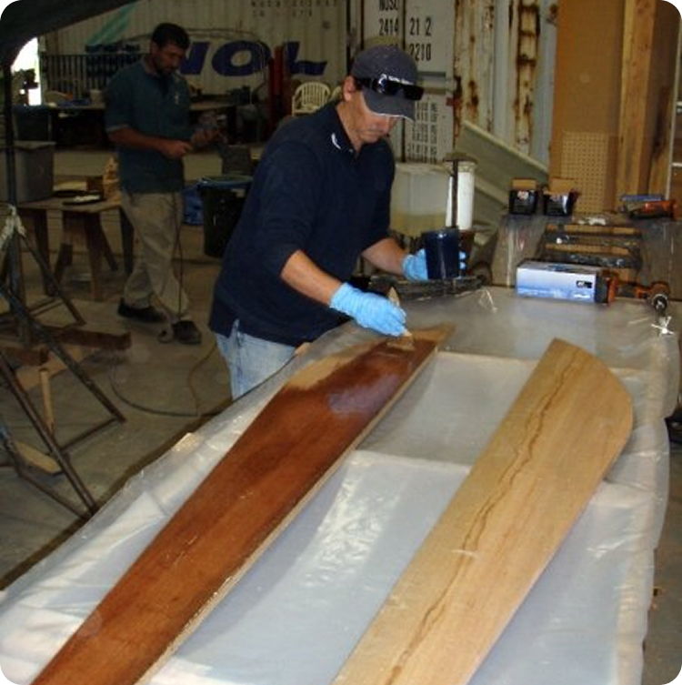 A man wearing gloves and sunglasses applies a finish to a long wooden board in a workshop, evoking the craftsmanship behind Schärenkreuzer, whilst another person works in the background amid scattered tools and materials.