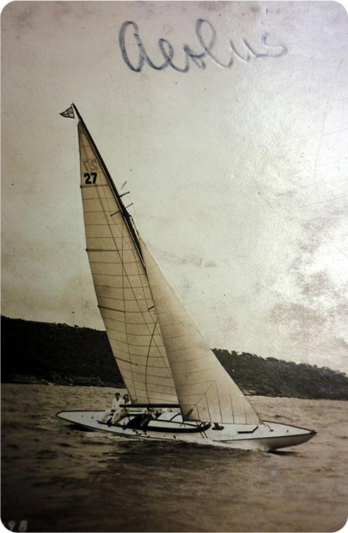 A vintage black-and-white photo of a Schärenkreuzer sailboat with the number 27 on its sail gliding on water. Two people are aboard, and Aeolus is handwritten at the top. A hilly shoreline is visible in the background.