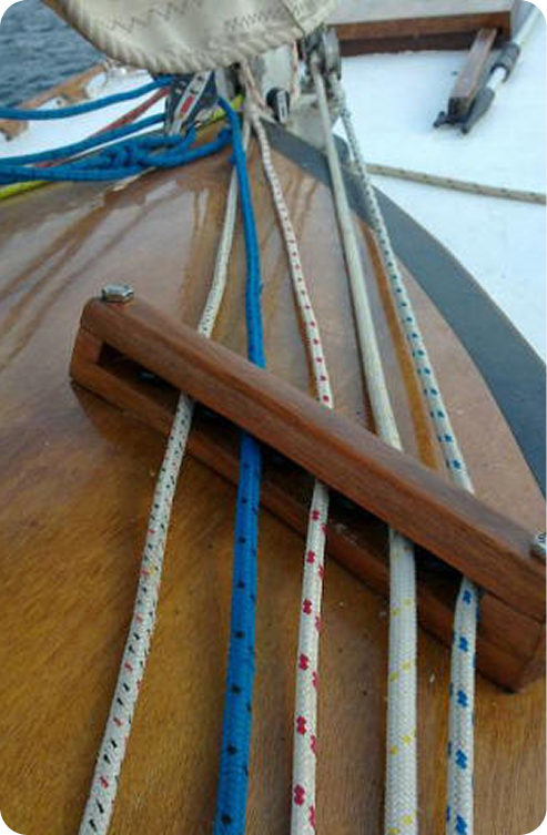 Close-up of a wooden Schärenkreuzer sailboat deck with colourful ropes threaded through a fairlead, part of the sail and mast visible in the background—a beautiful example of Classic Sailing Yachts craftsmanship.