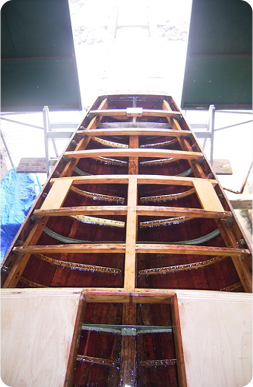 A wooden boat, reminiscent of Classic Sailing Yachts, is under construction inside a workshop, viewed from above with its exposed ribs and framework visible. Bright light pours in from large open doors at the far end.