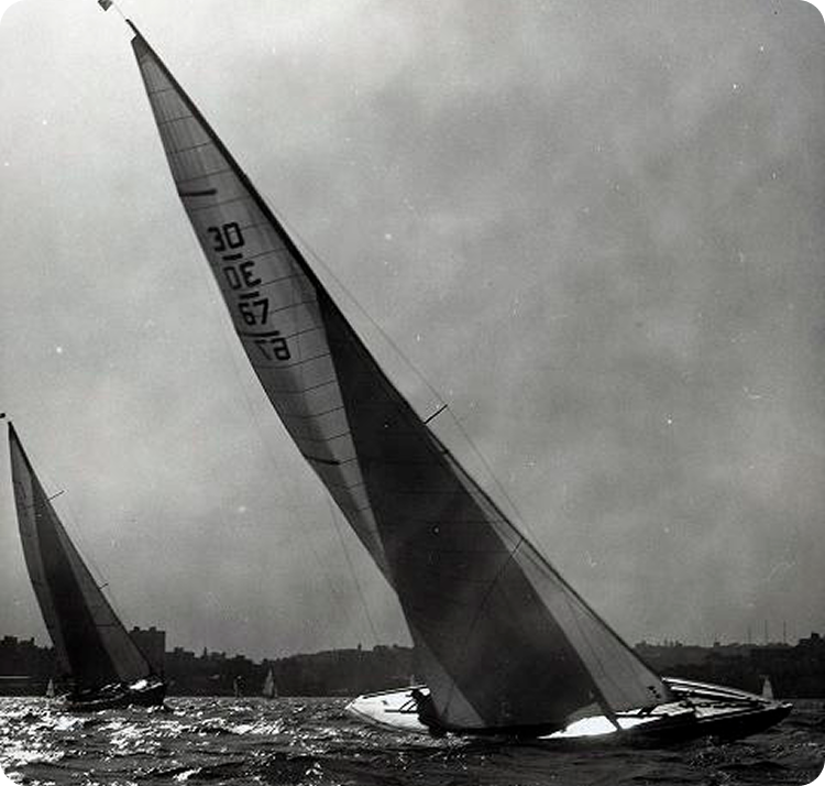 Black and white photo of two Skerry Cruisers with tall sails tilted on a choppy body of water, city shoreline visible in the background under a cloudy sky.