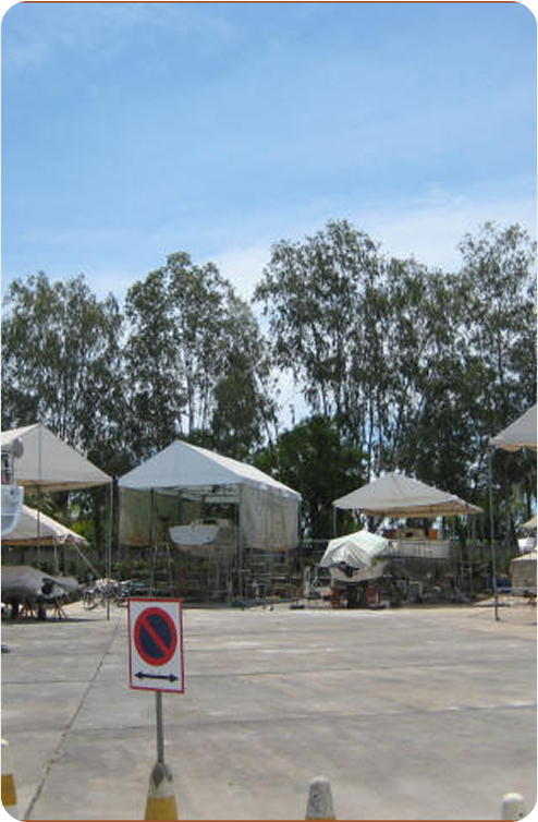 Several tents and structures are set up outdoors on a concrete surface, surrounded by tall trees. A “No Parking” sign is visible in the foreground. The sky is partly cloudy, evoking the atmosphere of a Classic Sailing Yachts gathering.