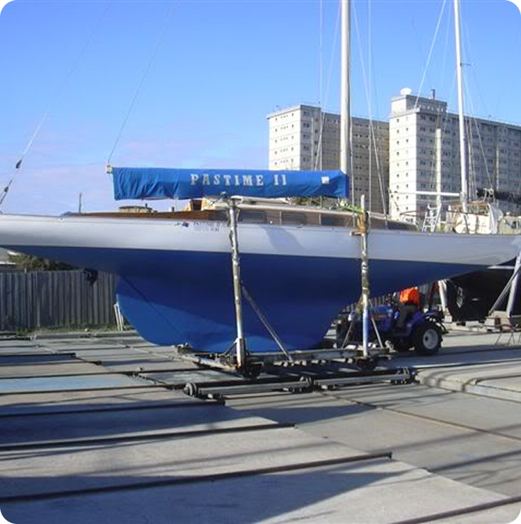 A white sailboat named Pastime II, a fine example of Classic Sailing Yachts, with a blue cover is mounted on a trailer at a marina, with tall buildings and other boats visible in the background.