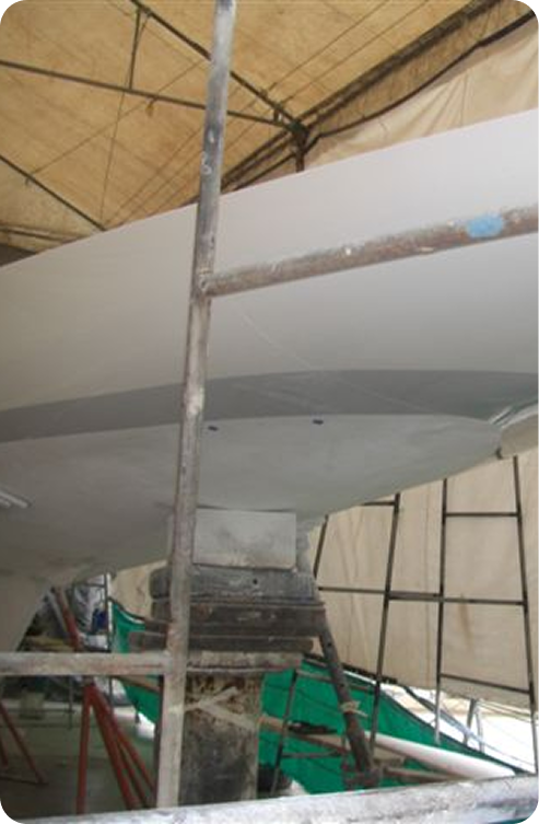 A close-up view of a Square Metre Yachts hull under construction inside a tented area, with scaffolding and support structures surrounding the classic sailing vessel.