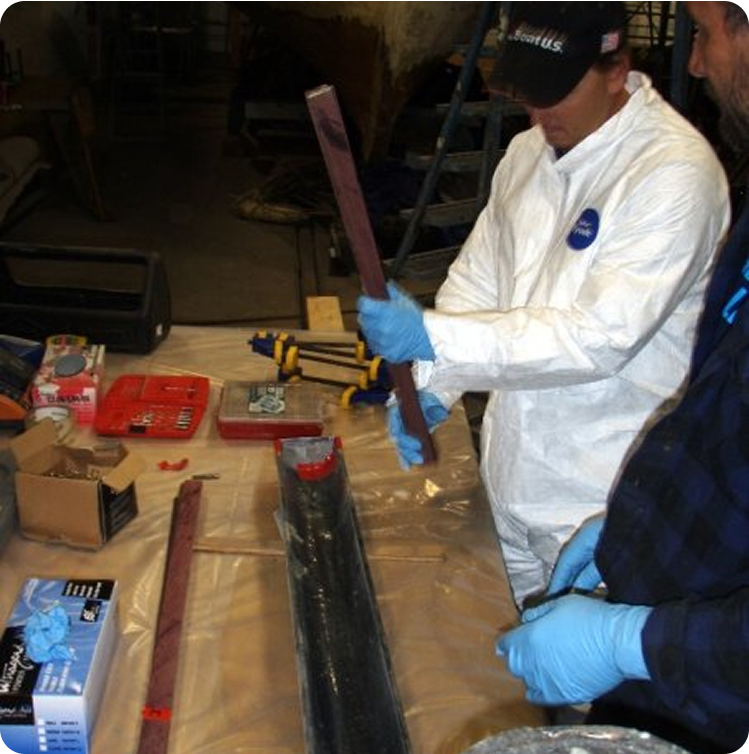 Two people wearing gloves and protective clothing work at a table covered in plastic, handling long rectangular objects—possibly components for Skerry Cruisers or Square Metre Yachts—with various tools and materials spread out before them.