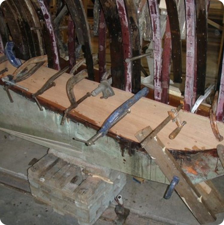 A wooden boat, resembling classic Schärenkreuzer or Square Metre Yachts, is under repair with new planks being clamped to its exposed ribs. Various tools and wooden blocks are scattered nearby amidst the restoration work.